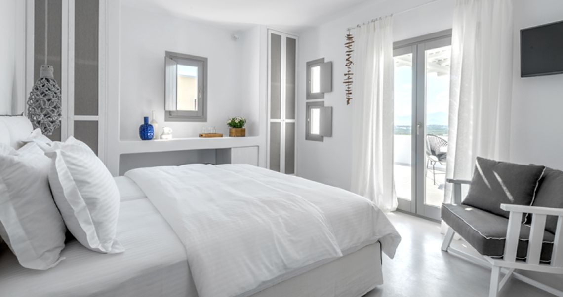 Spacious white bedroom suite featuring a king-sized bed, grey wardrobe panels, and double glass doors opening onto a sun-drenched private balcony with a view.