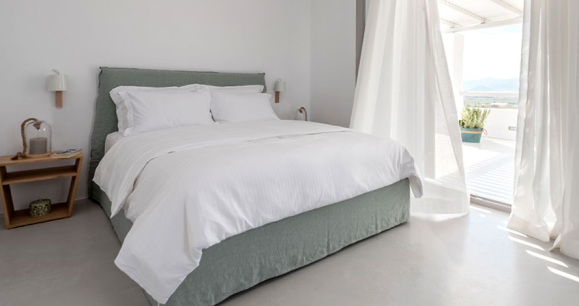 Serene bedroom with a sage green bed and white linens, featuring sheer curtains blowing in the breeze from the open balcony.