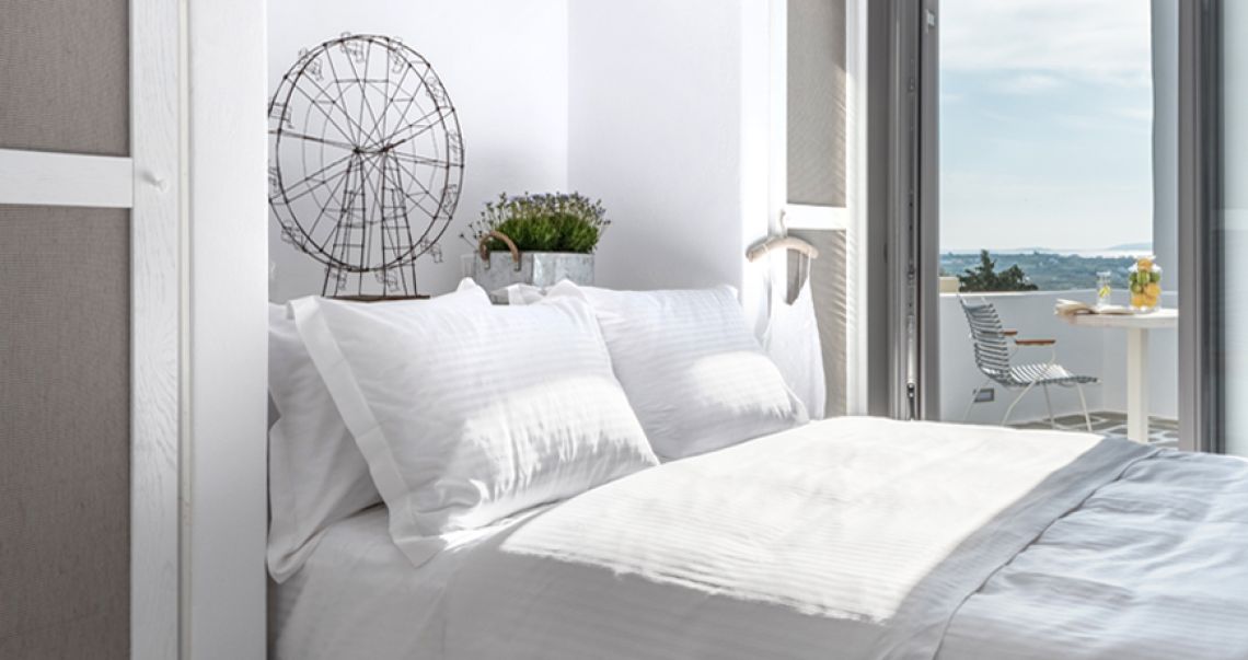 Sunlit bedroom with white linens and a wire Ferris wheel sculpture, opening onto a balcony with a table and sea view.