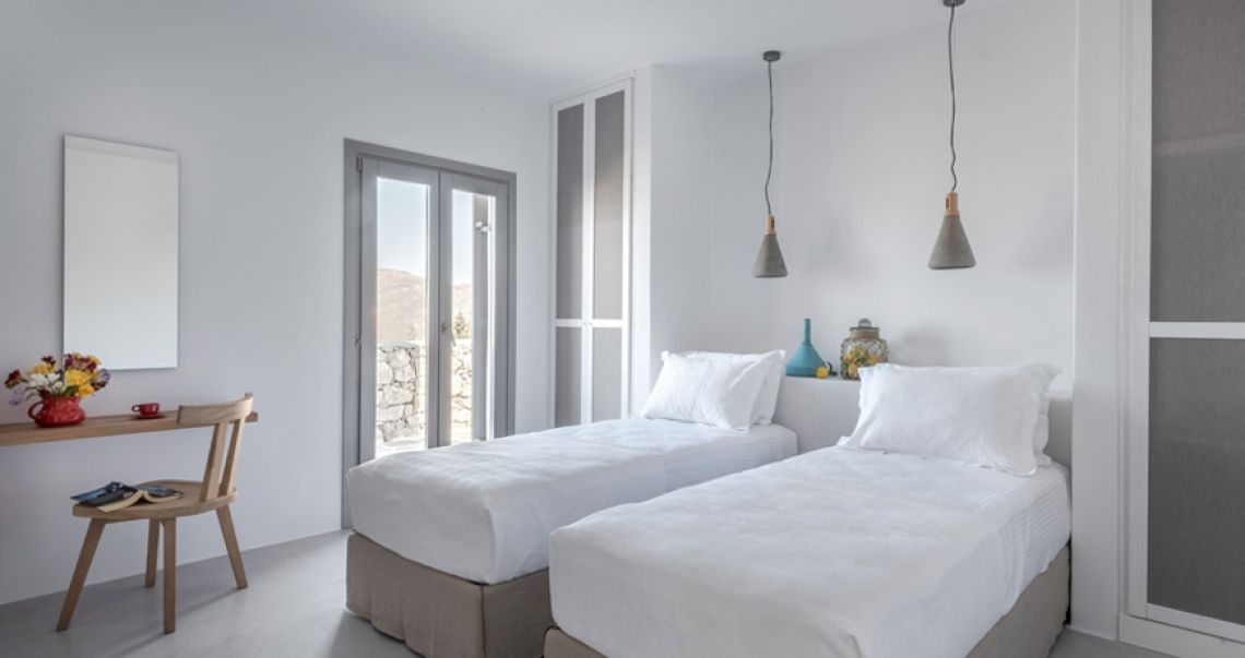 Bright, modern bedroom with two twin beds and tall grey wardrobes, featuring a small wooden desk area and glass doors leading to a balcony.