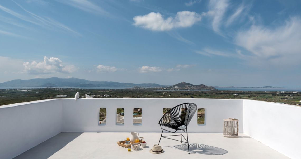 Open-air white balcony with a black Acapulco chair and breakfast tray, offering an expansive view of green fields and the sea.