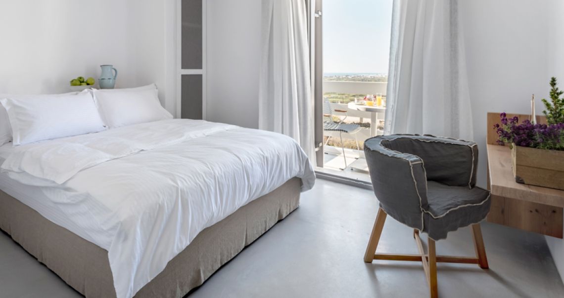 Bright, airy bedroom with a white bed, a grey linen armchair, and balcony doors opening up to a sunny landscape view.