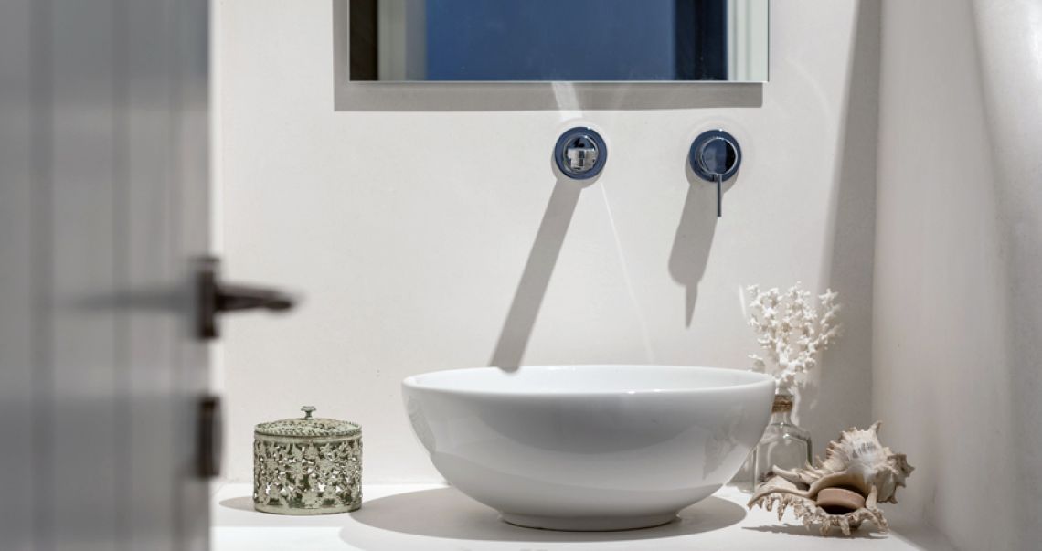 Minimalist bathroom vanity detail featuring a round white vessel sink, chrome wall-mounted faucets, and decorative white coral and seashells.