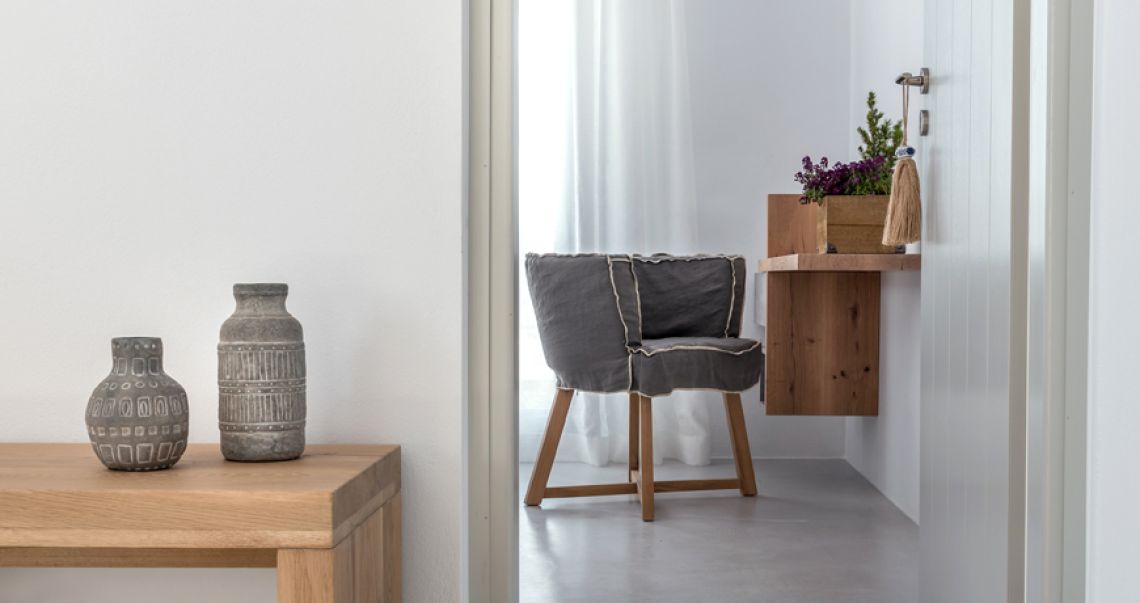 Interior detail showing a wooden console table with grey ceramic vases and a grey linen armchair in a minimalist white room.