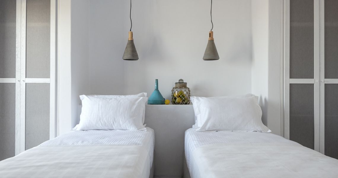 Symmetrical twin bedroom featuring white linens, hanging concrete pendant lights, and a central built-in shelf decorated with a blue funnel and a jar of lemons.