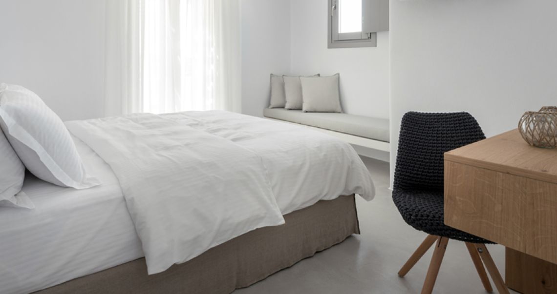 Bright white bedroom featuring a built-in window seat with grey cushions, a wooden desk, and a black woven chair.
