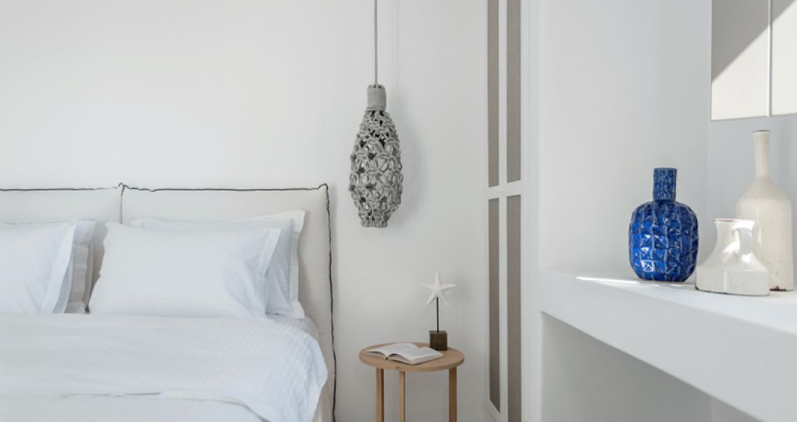 Minimalist white bedroom featuring a grey macrame pendant light hanging over a small wooden side table, and a built-in shelf displaying a blue textured vase.