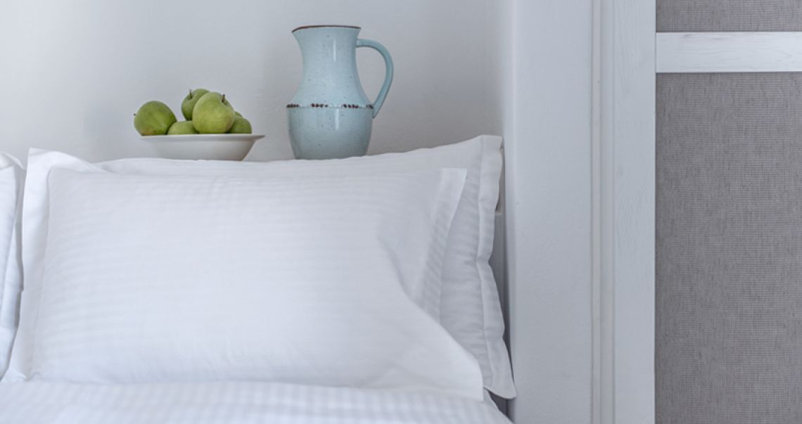Close-up detail of crisp white bed linens, accented by a bowl of green apples and a light blue pitcher on the headboard shelf.