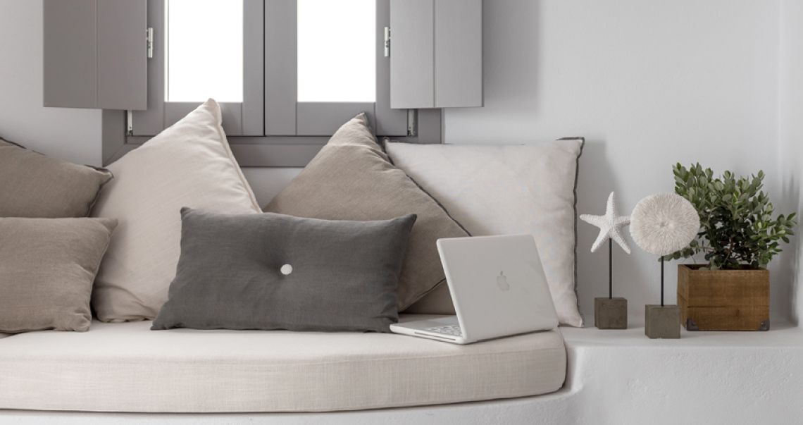 Close-up of a cozy window seat workspace with an open laptop, grey pillows, and decorative white coral pieces.