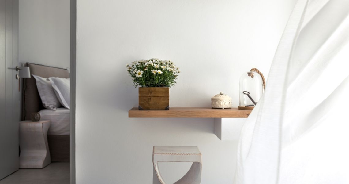 Detail of a floating wooden shelf in a bedroom holding a box of white daisies and a glass cloche, next to a breezy white curtain.