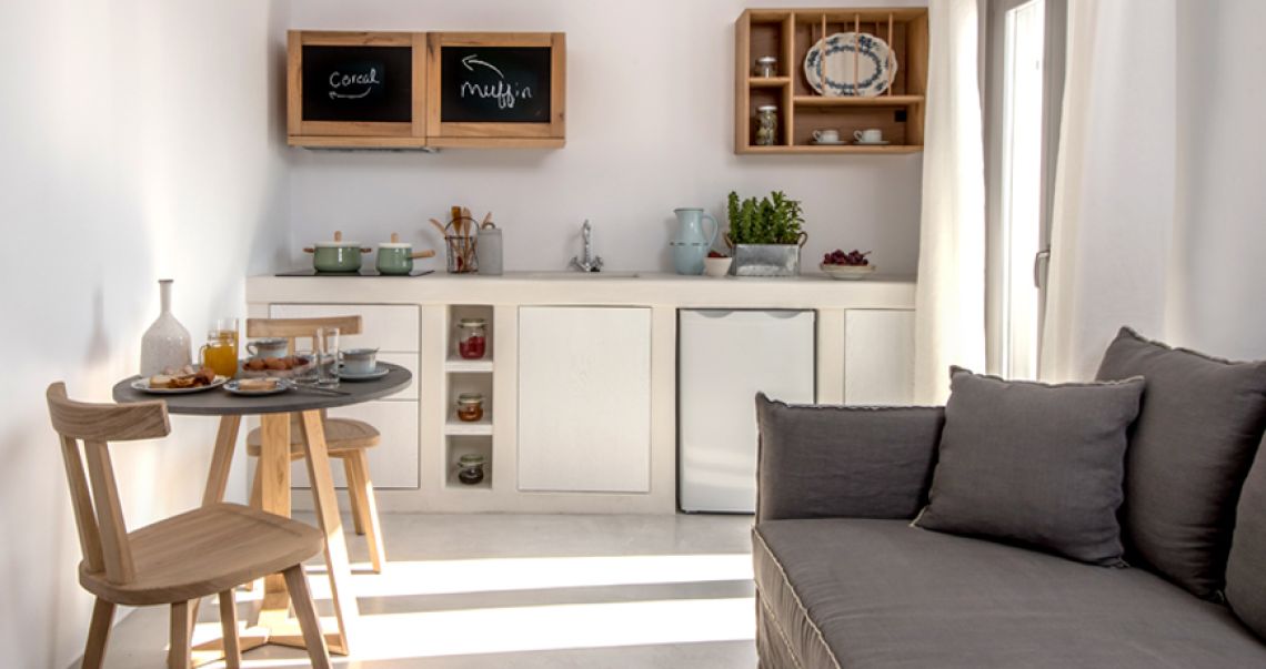 Bright open-plan kitchenette and living area featuring white built-in cabinets, a small round wooden dining table, and a grey sofa.