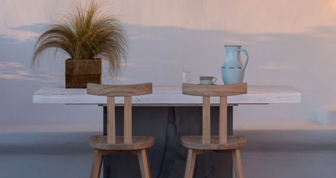 Intimate outdoor dining setup at dusk featuring a rustic white wooden table, two wooden chairs, and a blue pitcher against a wall illuminated by soft sunset light.