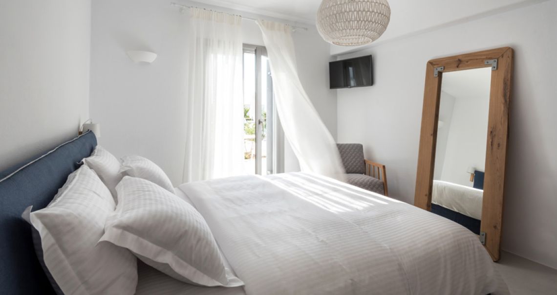 Bright and breezy bedroom featuring a large rustic wooden floor mirror, a blue headboard, and sheer curtains billowing from the open balcony door.
