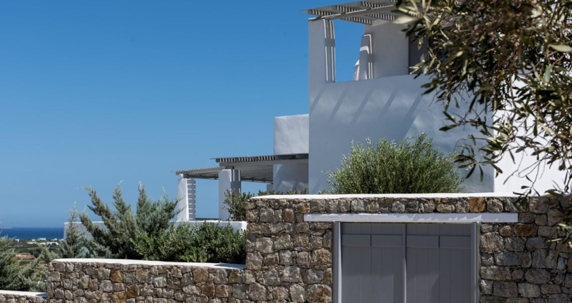 Multi-level white villas with pergolas and stone walls, framed by olive branches against a bright blue sky.