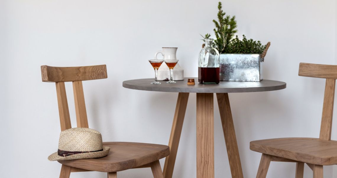 Intimate dining setup featuring a round wooden table with aperitifs, a glass decanter, and a straw hat resting on a chair.