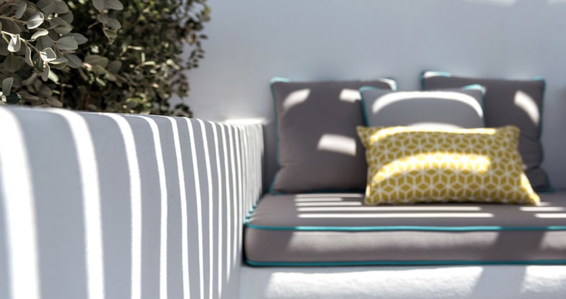 Close-up of outdoor built-in seating with grey cushions and a bright yellow geometric throw pillow, cast with artistic striped shadows from the pergola above.