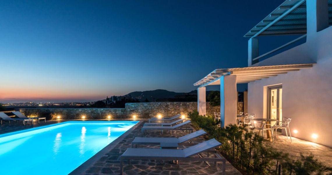 Illuminated outdoor swimming pool and patio area at dusk, featuring warm ambient lighting and a sunset gradient sky.