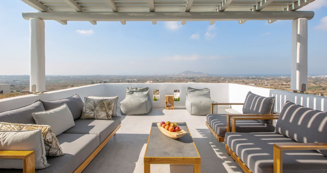 Spacious rooftop terrace featuring grey sofas, bean bag chairs, and a wooden coffee table with fruit, overlooking a panoramic landscape.