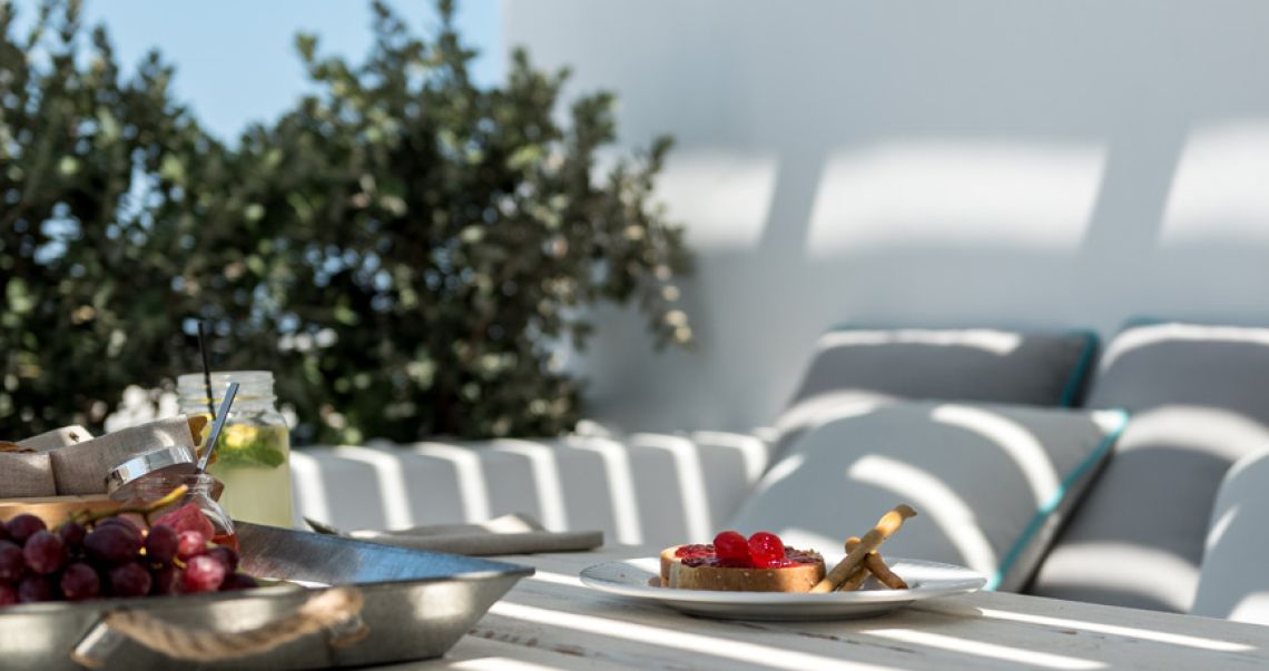 Close-up of a sunny terrace breakfast featuring toast with red jam, fresh grapes on a metal tray, and jarred lemonade, framed by green foliage.