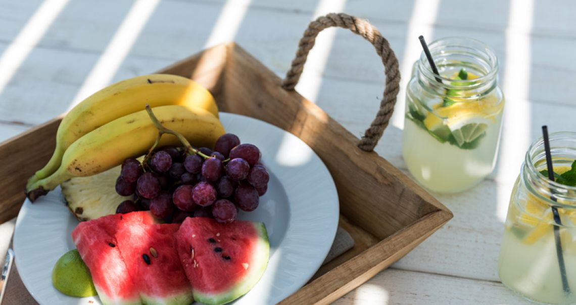 Close-up of a fresh fruit platter with watermelon, grapes, and bananas served on a wooden tray next to jarred lemonade.